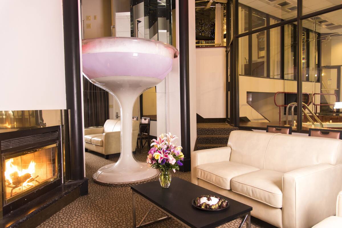 Un hall d’hôtel moderne avec une cheminée, un canapé blanc, une table basse noire ornée d’un vase de fleurs et un grand objet décoratif en verre.