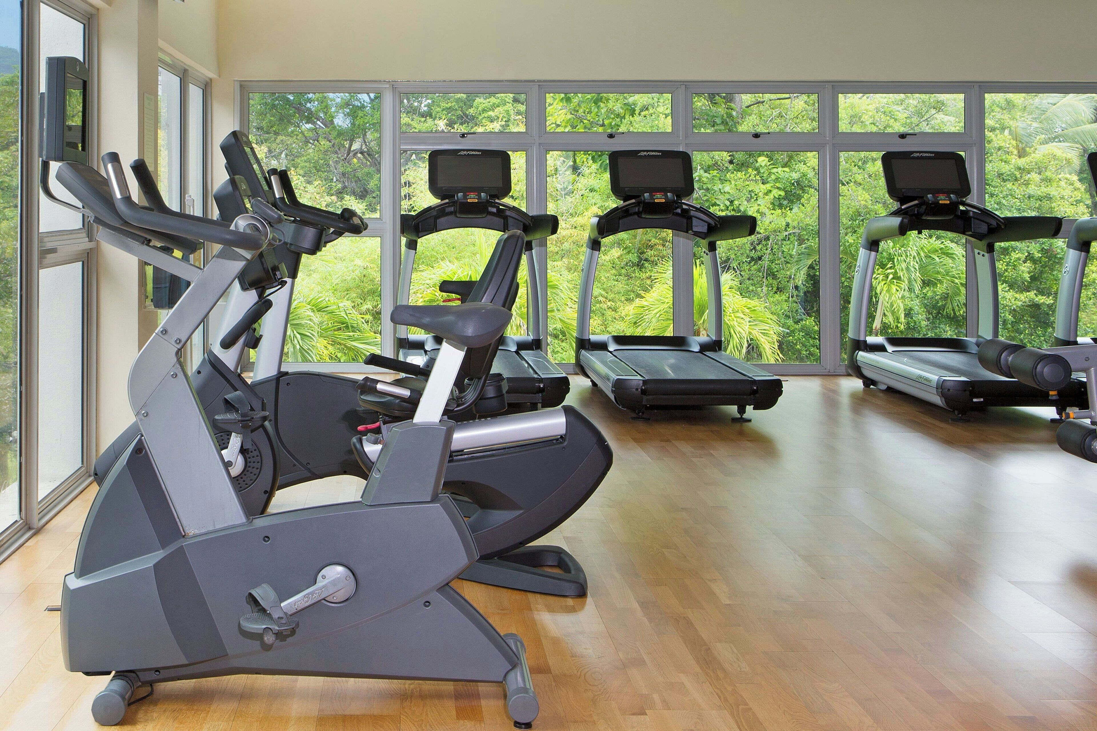 Une salle de sport équipée de vélos d’exercice et de tapis de course, avec de grandes fenêtres donnant sur un espace verdoyant.