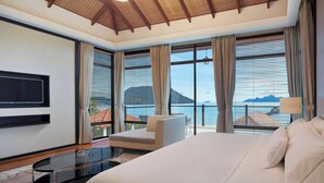 A modern hotel room with a large bed, a TV mounted on the wall, a seating area with a chair, and large windows offering a view of the sea and buildings.