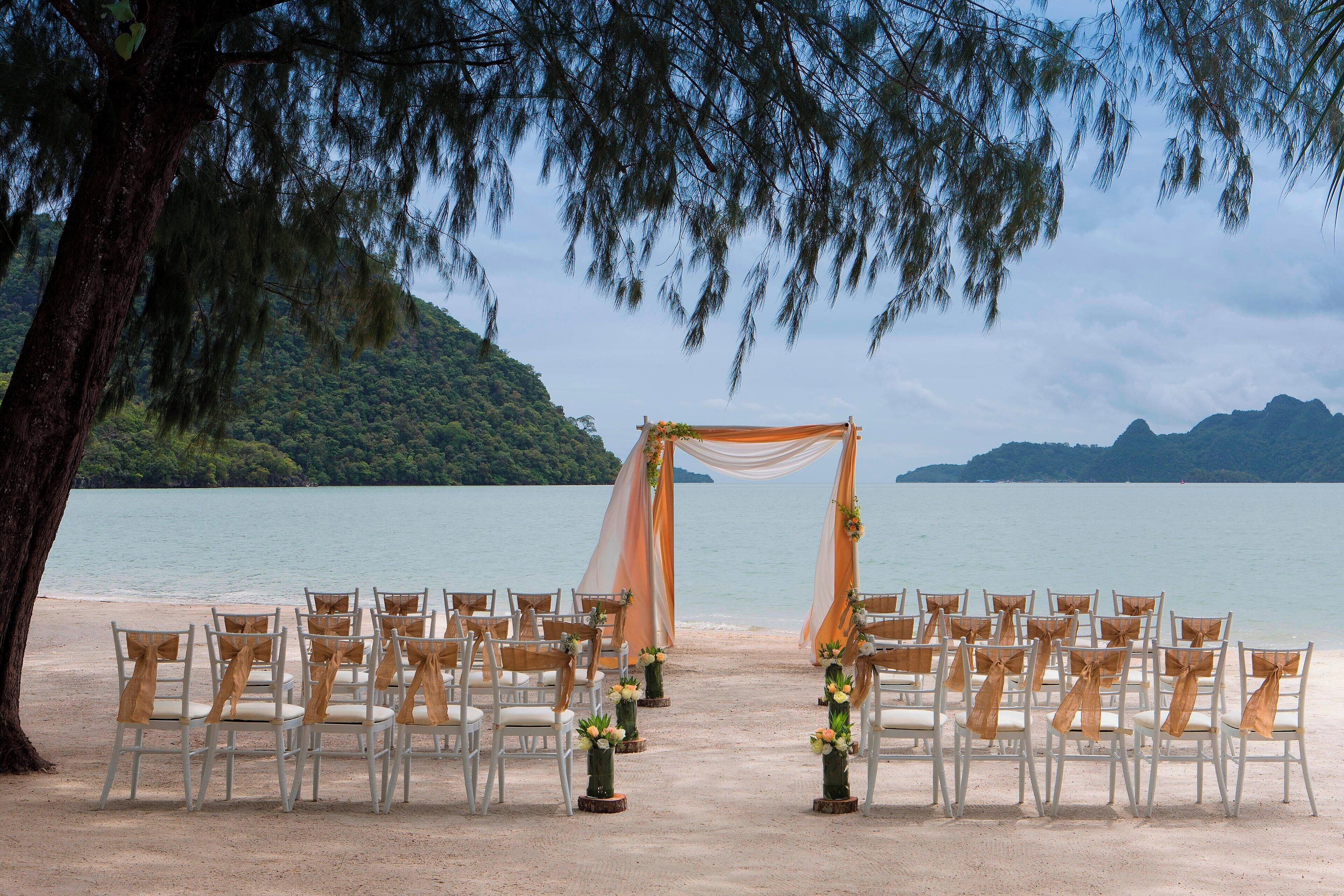 A beach wedding setup with chairs arranged in rows, a canopy, and a backdrop of a calm sea and hills.