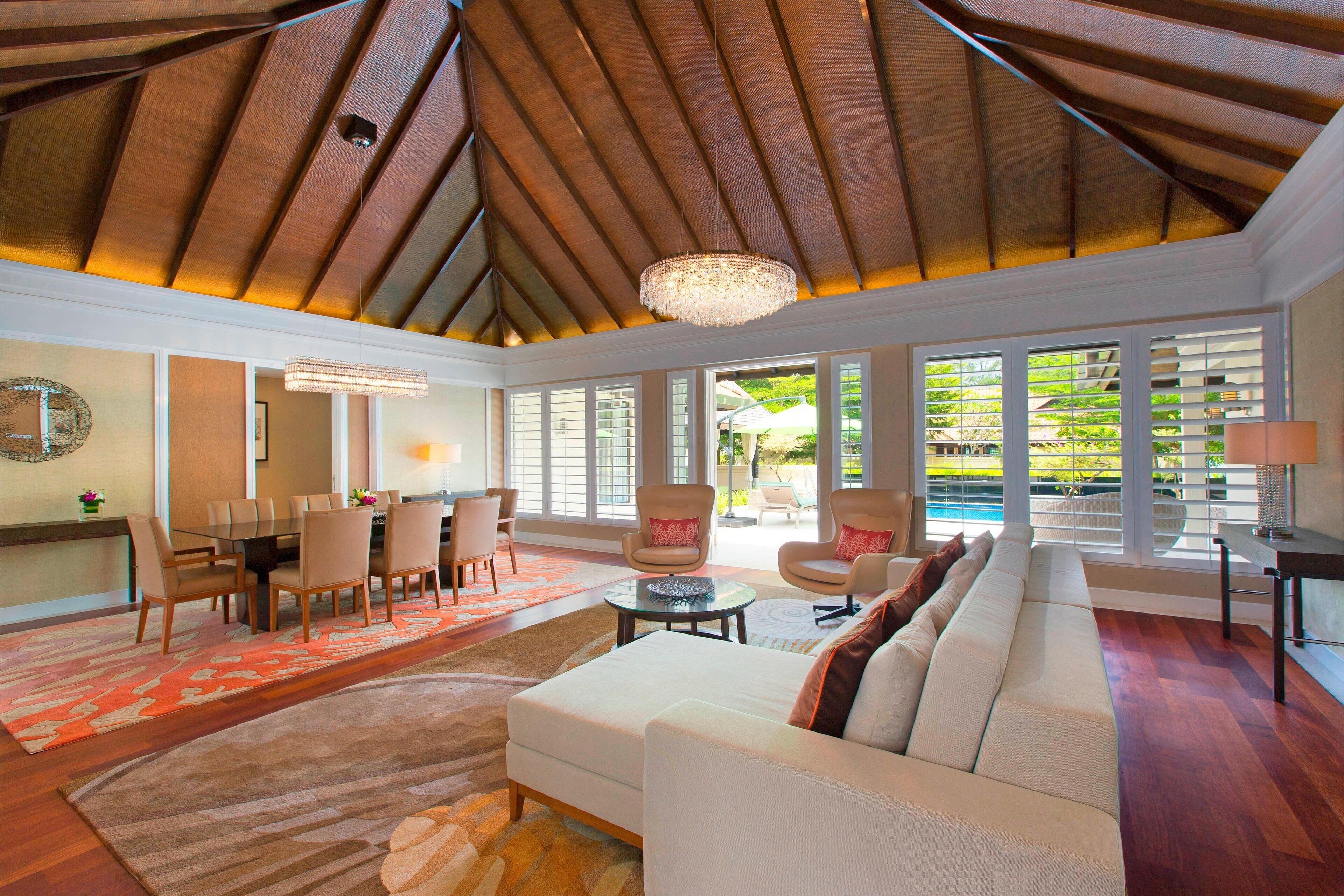 A spacious living area with a high ceiling, a large sofa, a dining table, and a view of a pool through large windows.