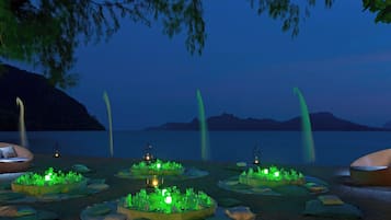 A beachside dining setup with tables and chairs, illuminated by green lights, and a backdrop of a calm lake and mountains.