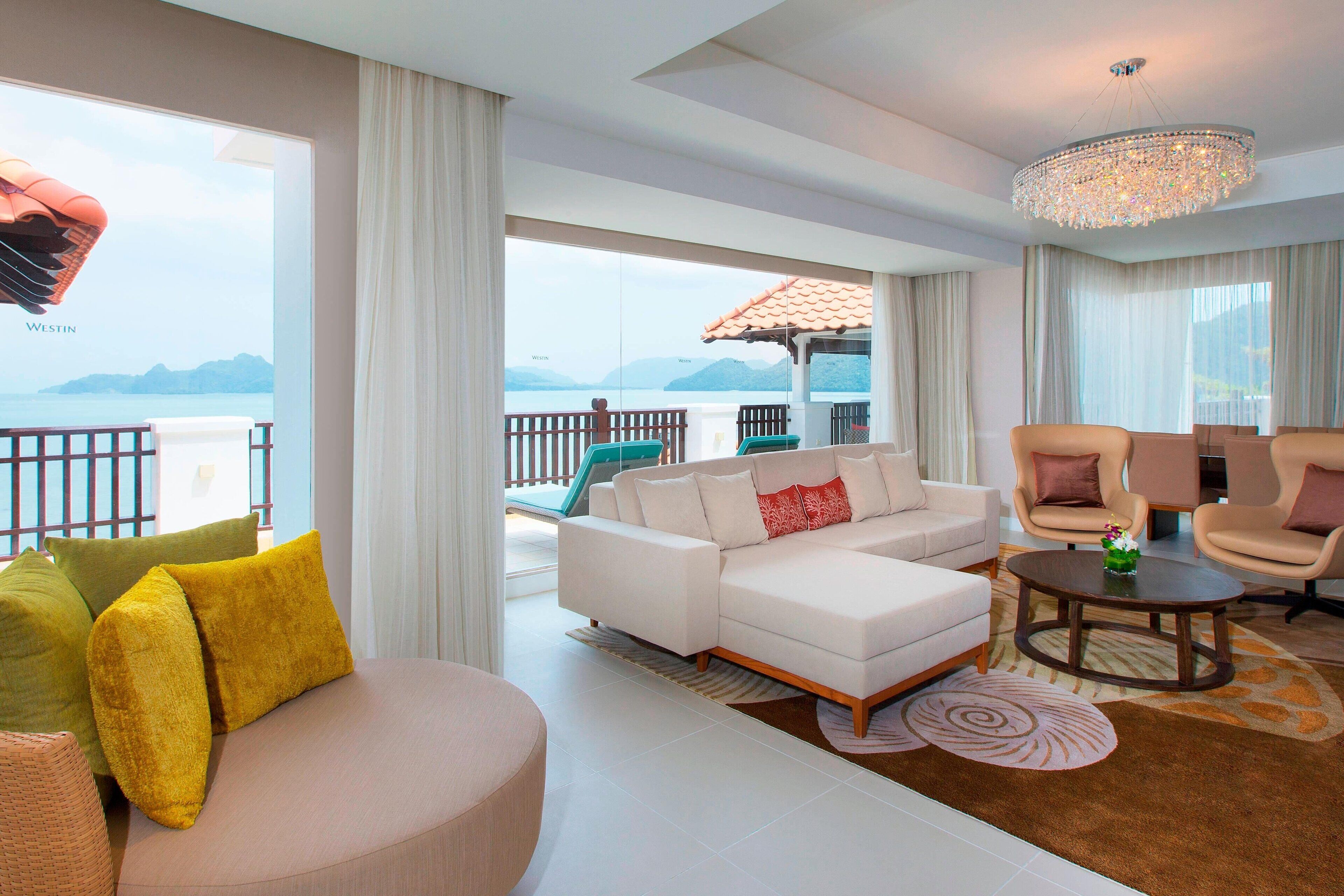 A modern living room with a white sofa, beige armchairs, and a round coffee table. A large window offers a view of the sea and mountains.
