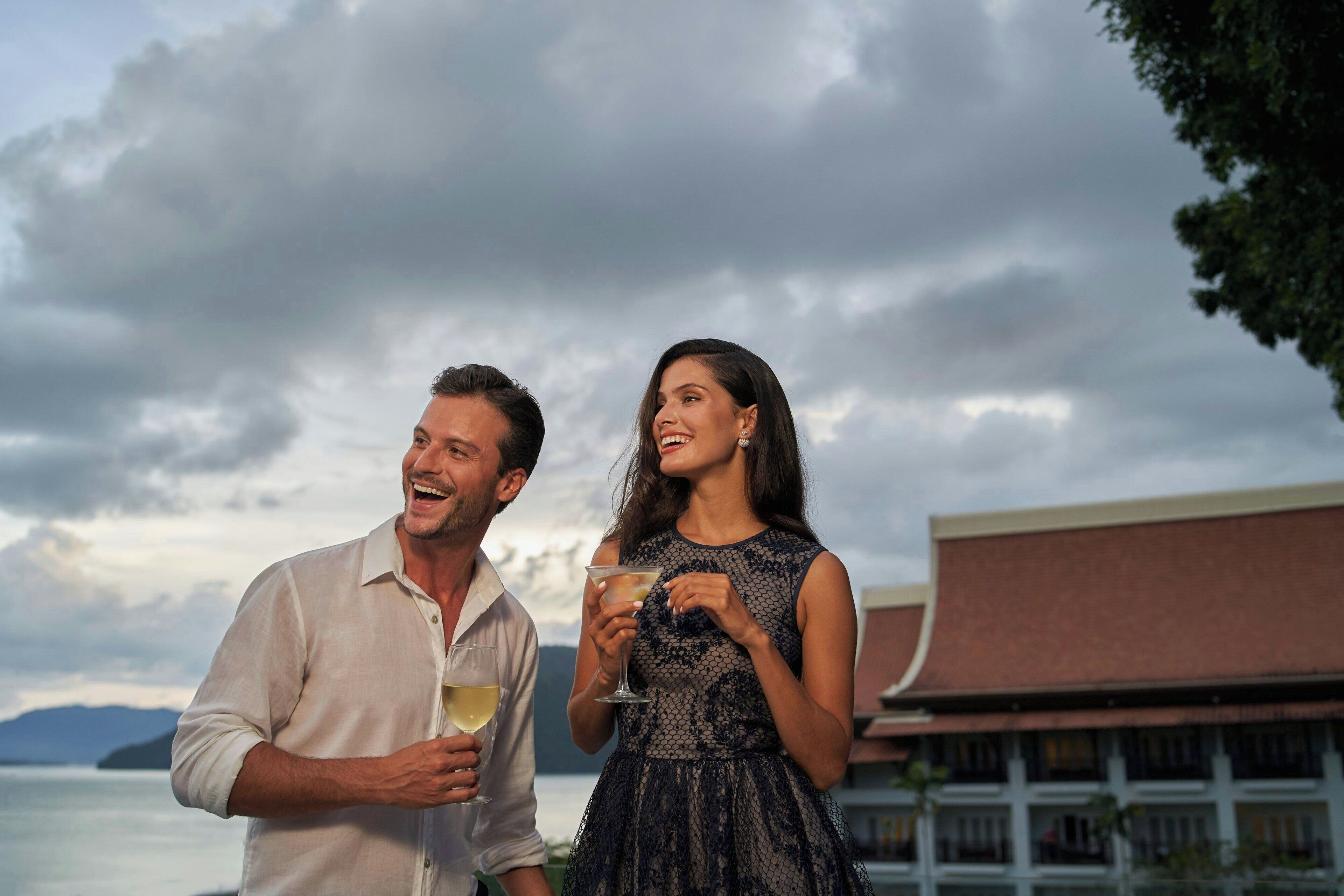 A couple standing outdoors with drinks, overlooking a body of water and a building with a red roof.