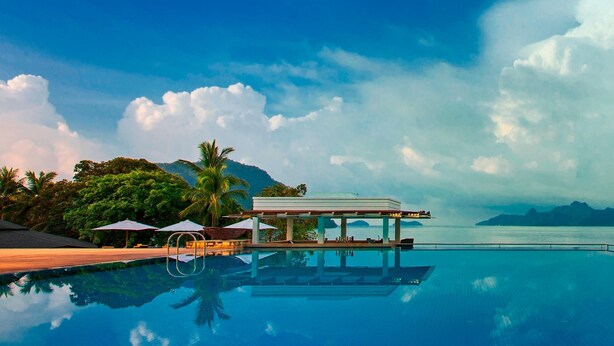 A poolside area with a covered seating area, palm trees, and a view of the sea.