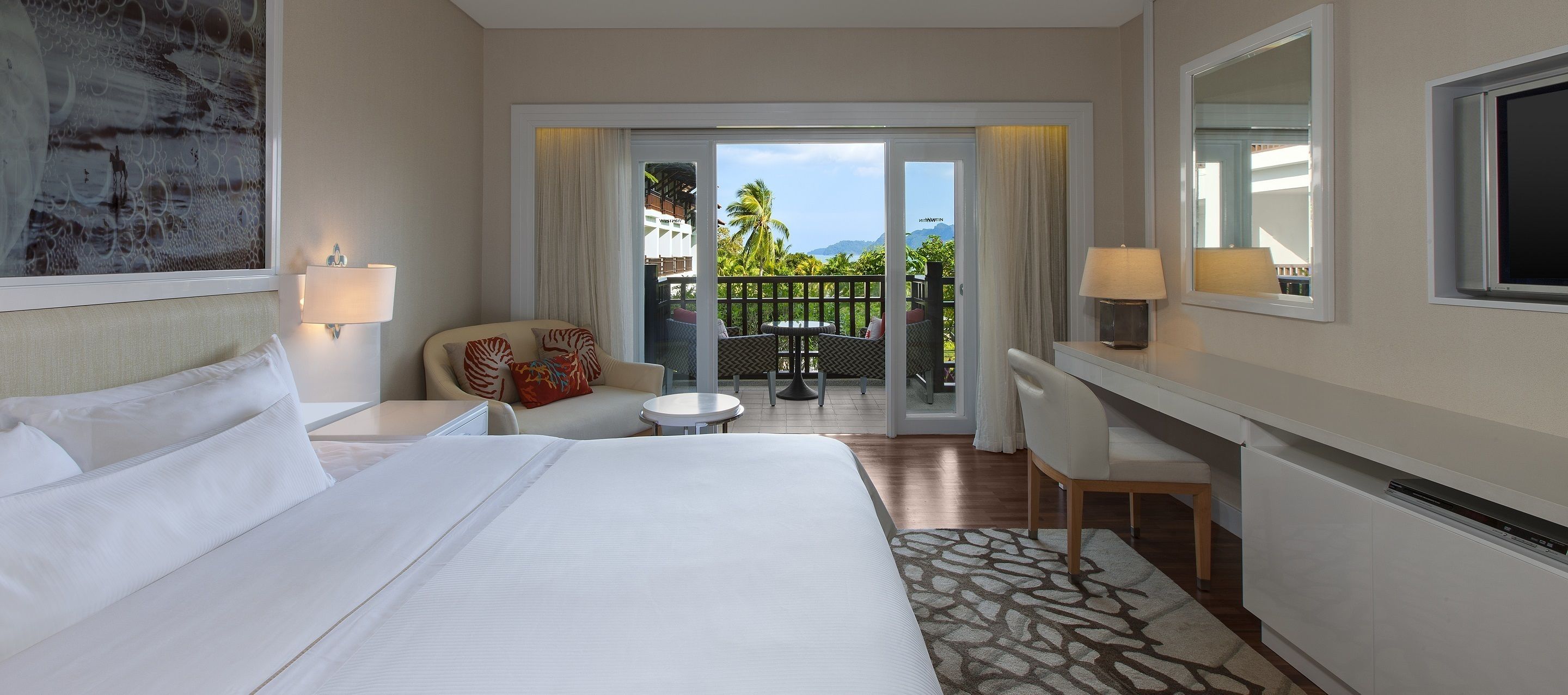 A hotel room with a large bed, a desk with a chair, a small round table, and a balcony with a view of palm trees and mountains.