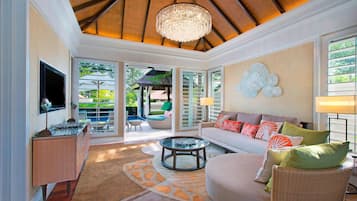 A spacious living room with a large chandelier, a sofa with colorful cushions, a glass coffee table, and a television on a wooden cabinet.