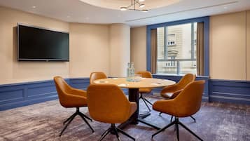 A conference room with a round table, four chairs, a flat-screen TV, and a large window with a city view.