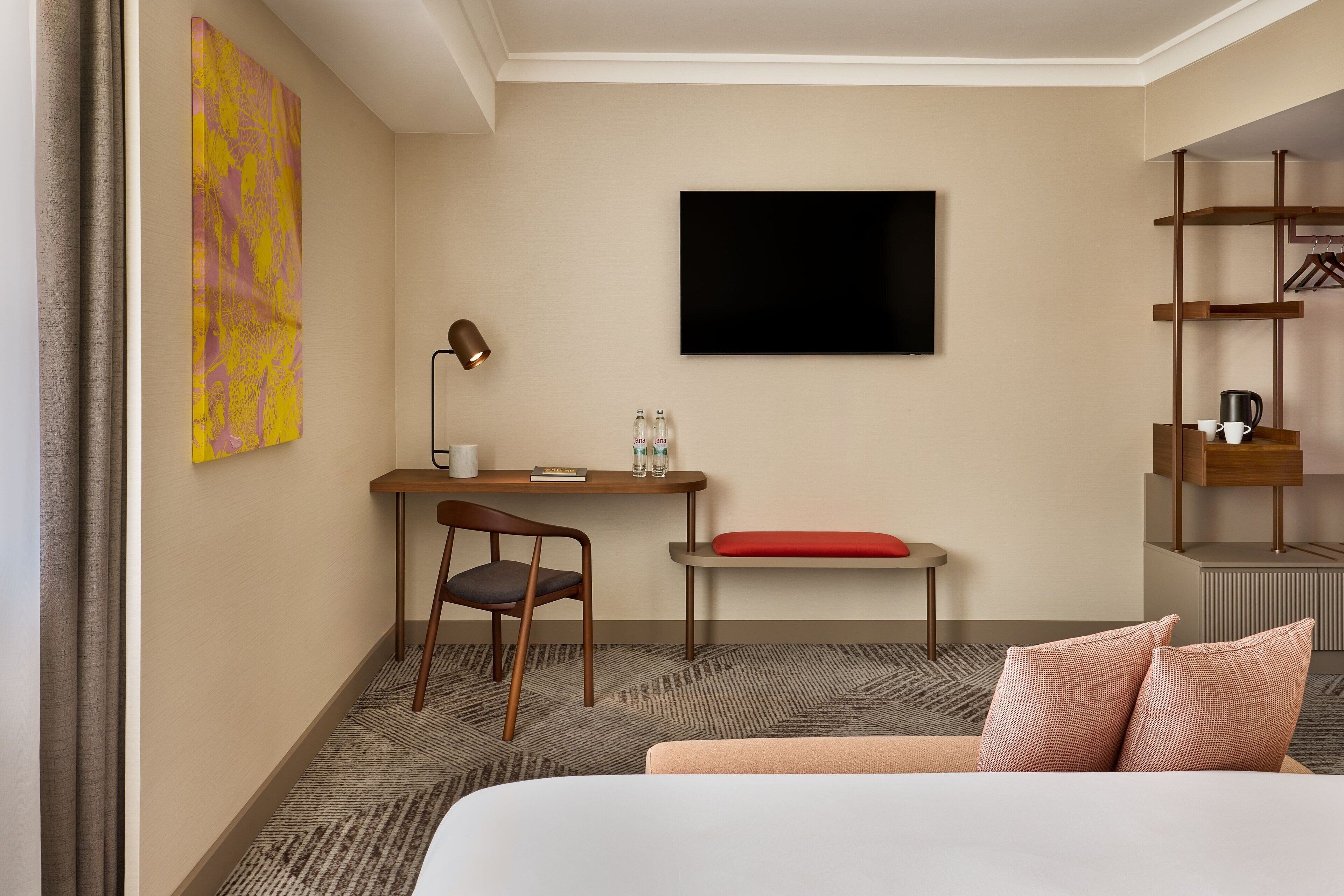 Una habitación de hotel con cama, escritorio con silla, televisor montado en la pared y repisa con cafetera.