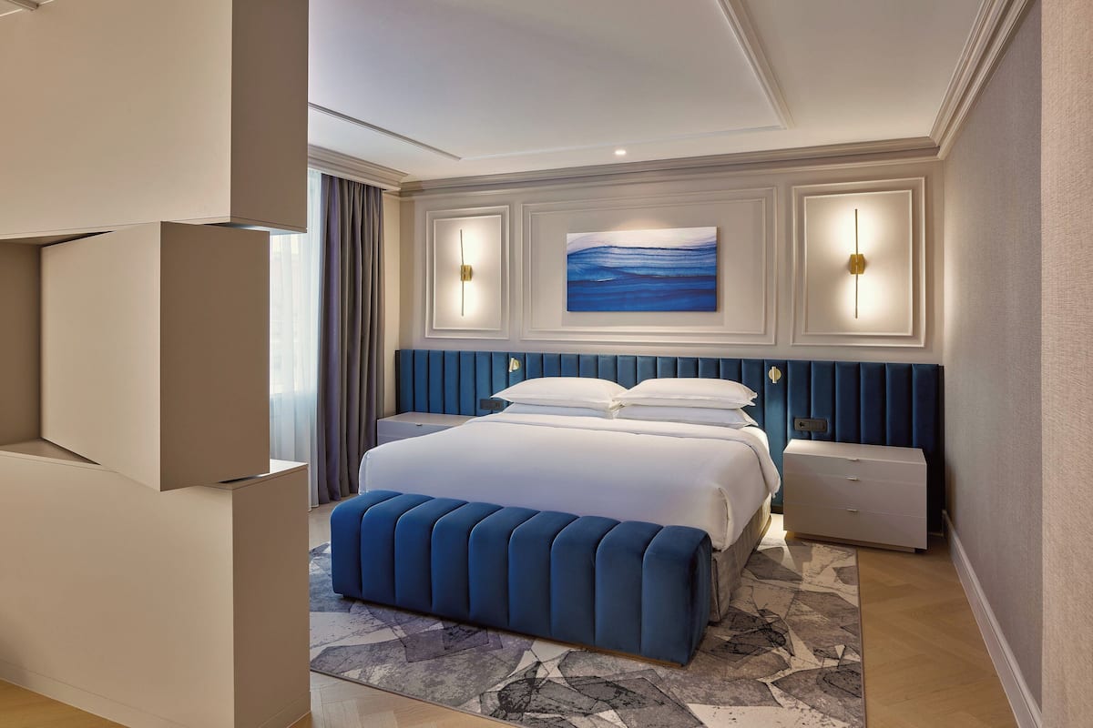 A modern hotel room with a large bed, a blue upholstered bench, a nightstand, and a painting above the bed.