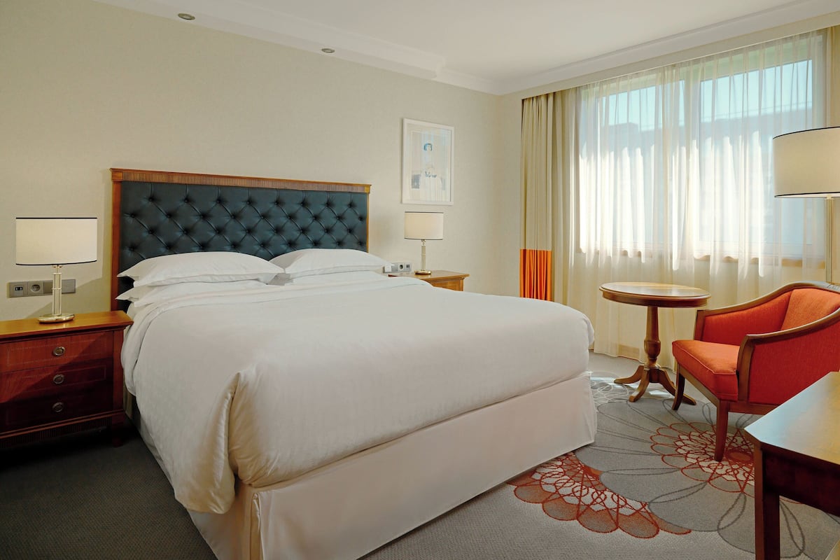 A hotel room with a large bed, a wooden nightstand, a red armchair, a small round table, and a window with sheer curtains.