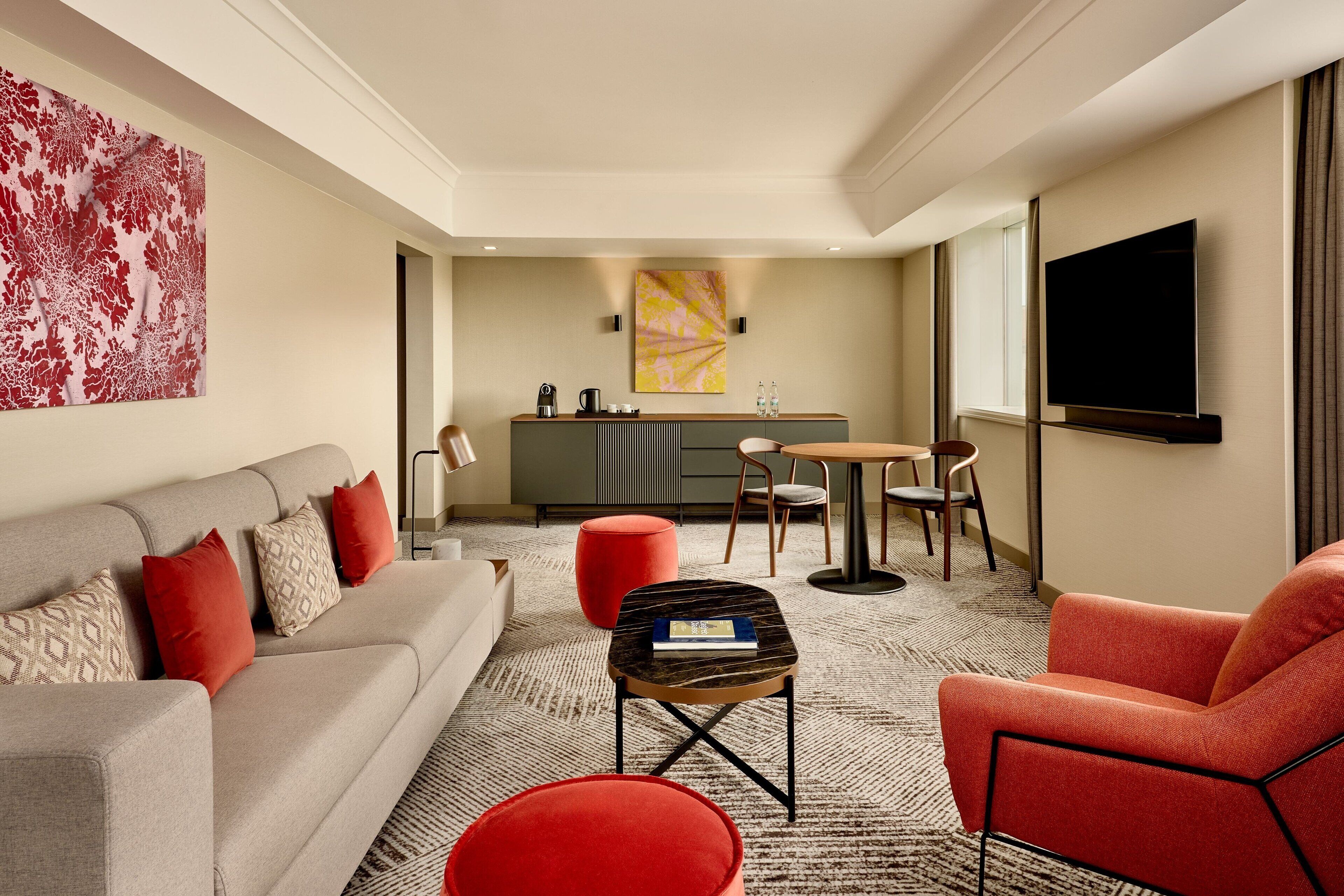 Una habitación de hotel moderna con sofá, un reposapiés rojo, una mesita, un televisor y una zona de cocina con mesa y sillas.