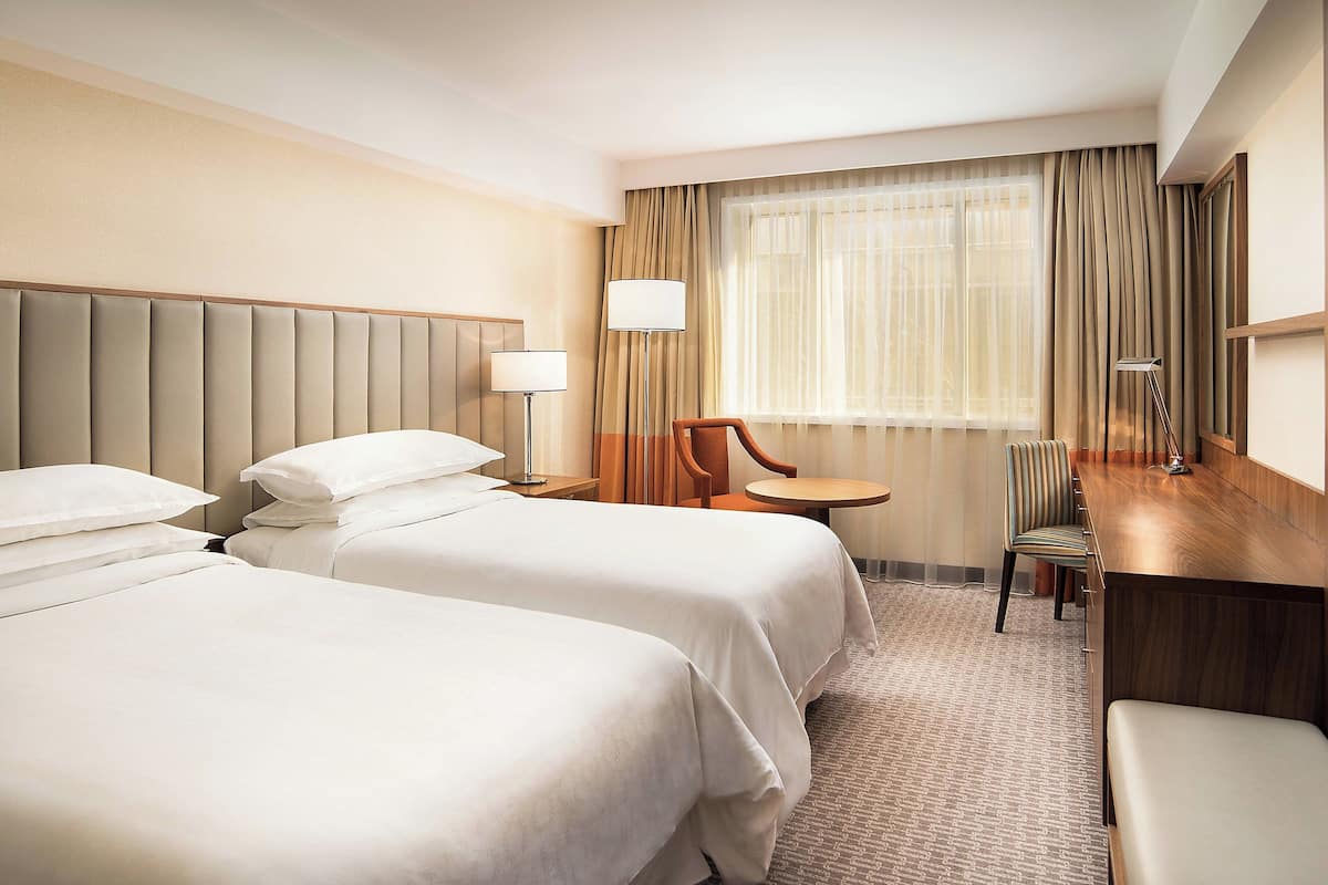 A hotel room with two beds, a desk, a chair, a lamp, and a window with curtains.