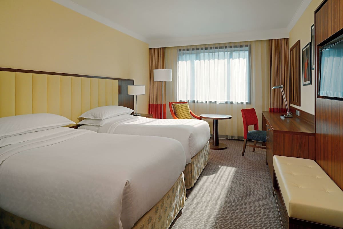 A hotel room with two beds, a desk, a chair, a lamp, and a large window with curtains.