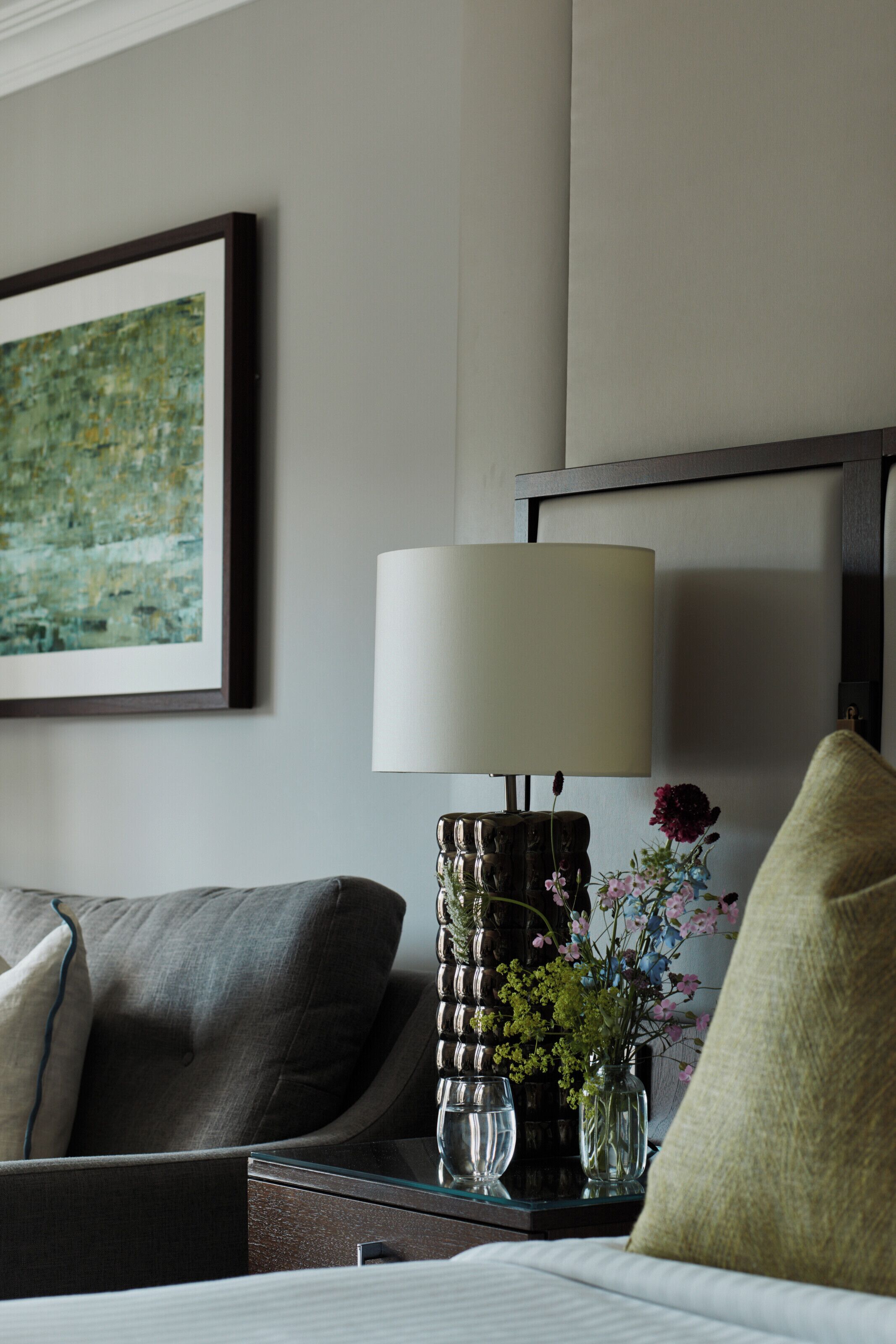 A hotel room with a bed, a nightstand with a lamp, a vase with flowers, a framed painting, and a dark-colored headboard.