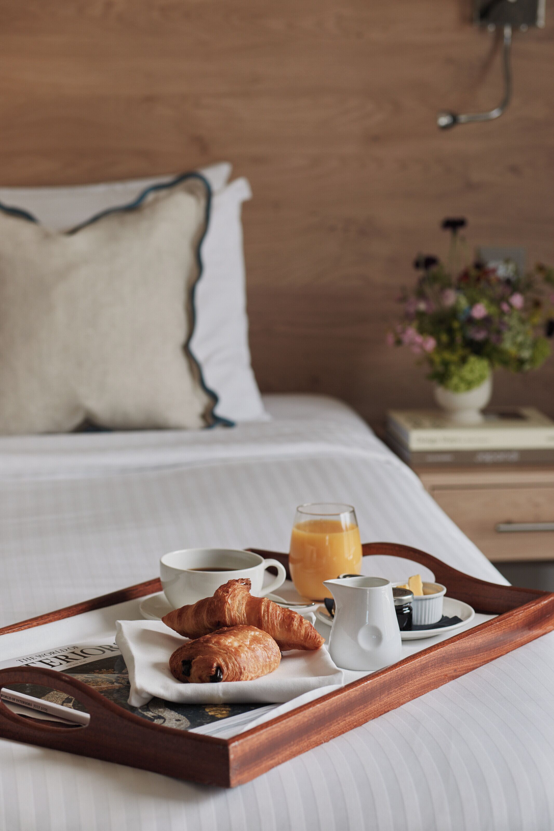 Bandeja de desayuno con cruasanes, café y jugo sobre una cama con almohada y mesita de noche con una planta.