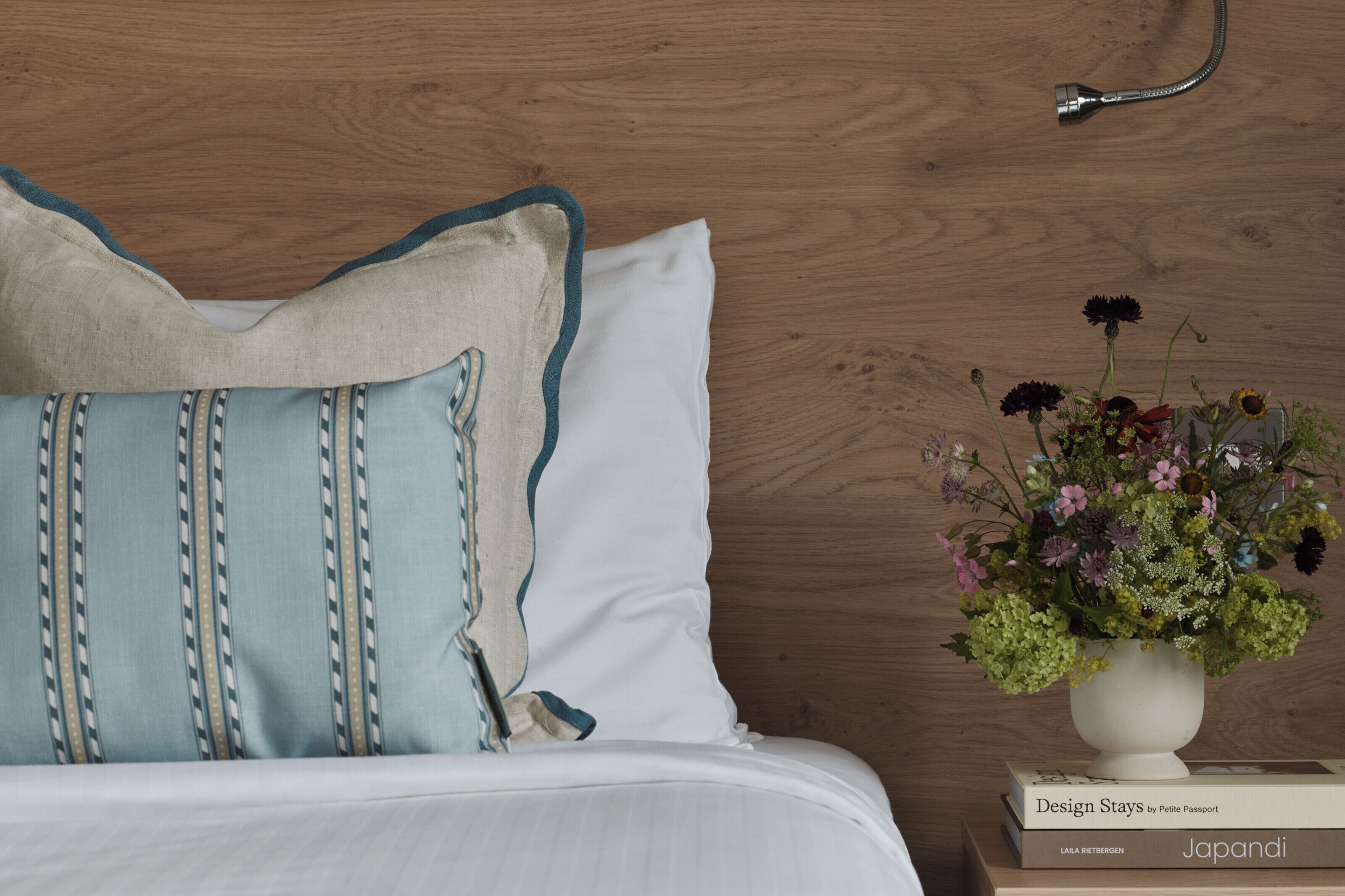 Una cama bien hecha con almohadas, un florero blanco con flores y un libro titulado 'Design Stays' sobre una mesita de noche de madera.