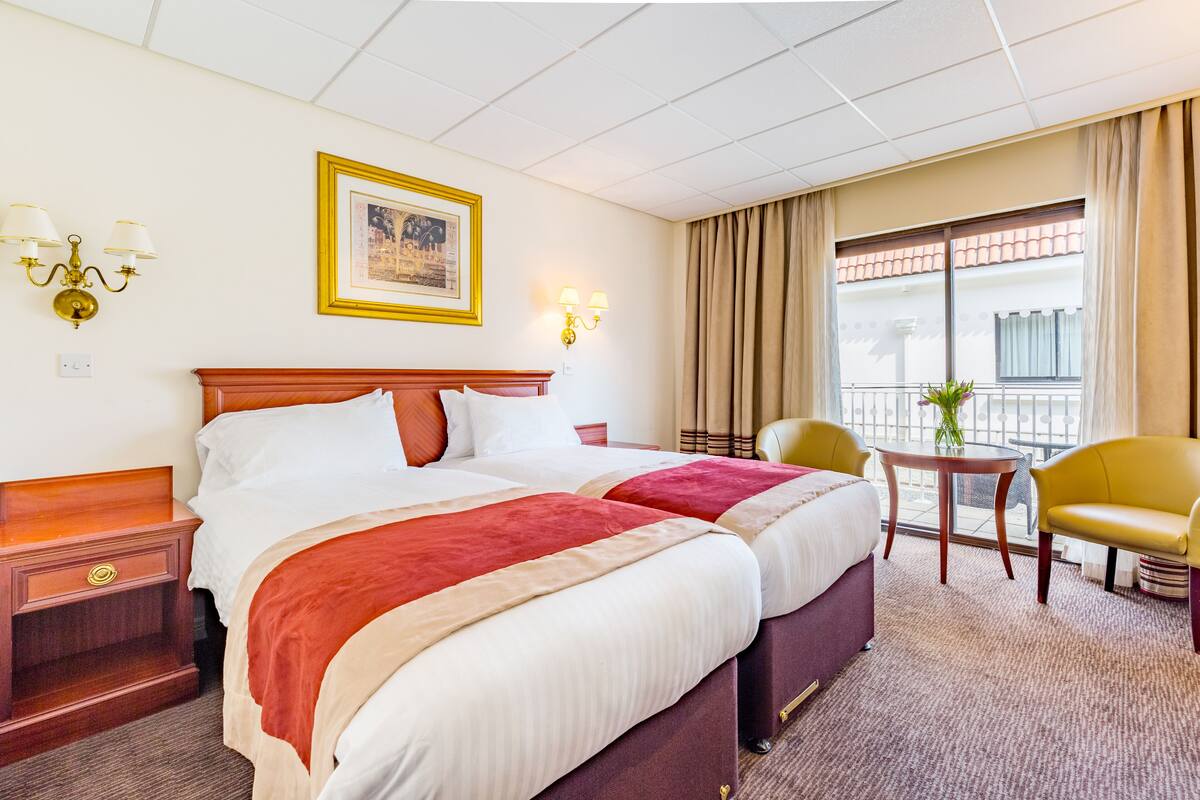 A hotel room with two beds, a wooden nightstand, a small table with a vase, a yellow armchair, and a balcony with a view.