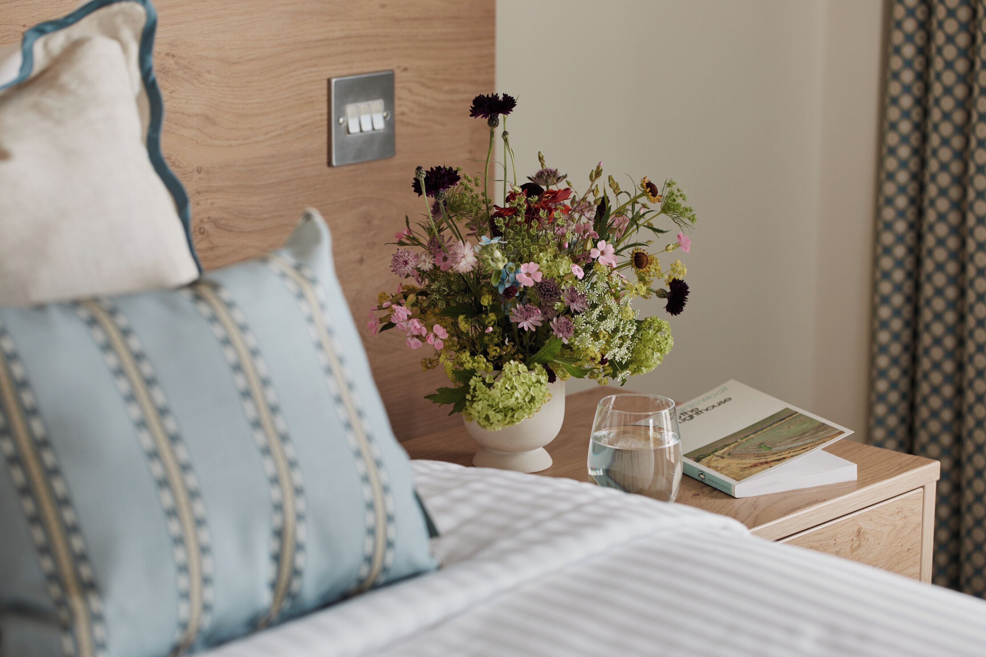 A neatly made bed with a pillow, a bedside table with a book and a glass of water, and a floral arrangement.