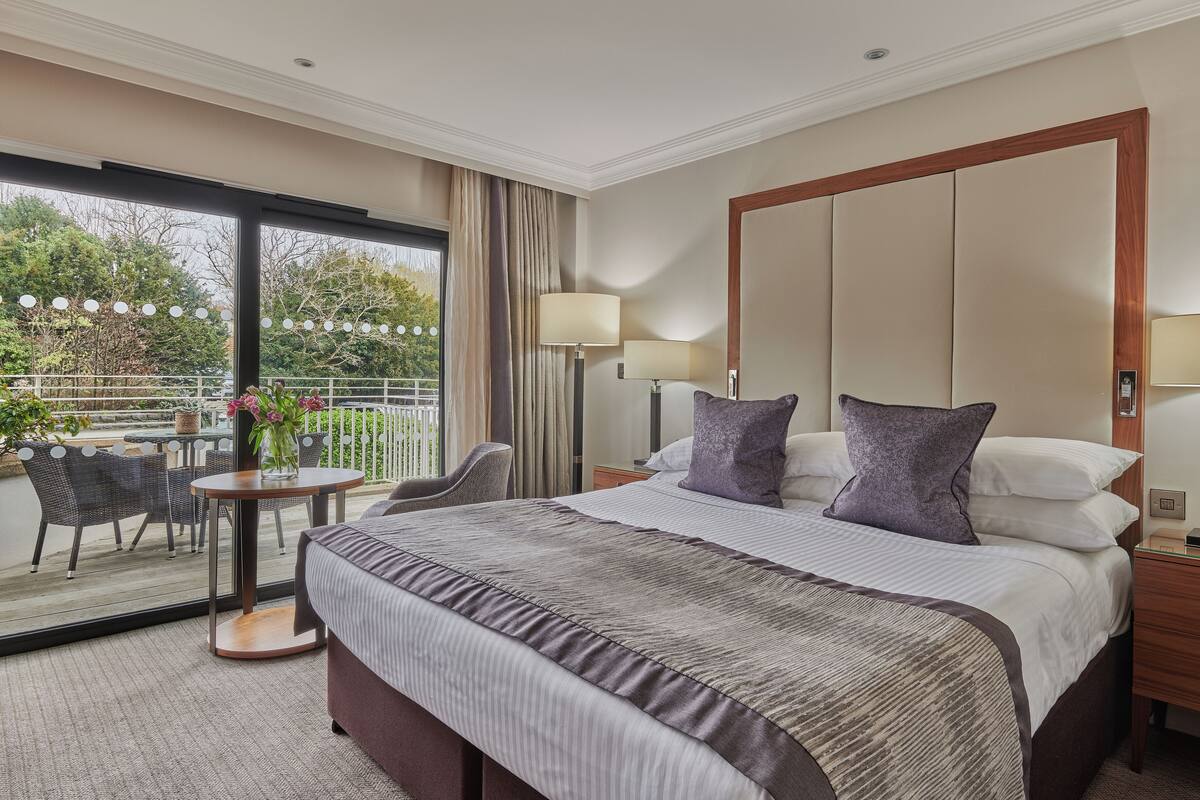 Quarto de hotel com uma cama grande, uma mesinha de cabeceira, uma luminária e vista para o exterior por uma porta de correr de vidro.