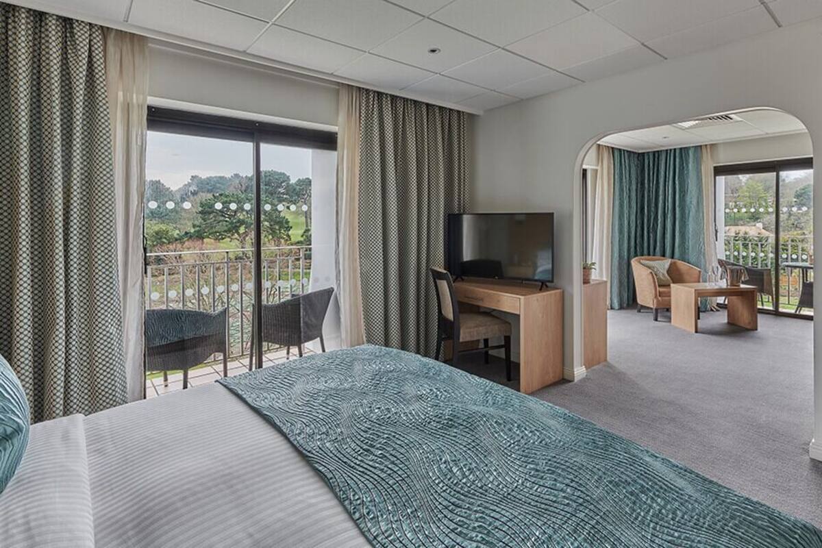 A hotel room with a bed, a desk with a TV, a balcony with chairs, and a view of greenery.