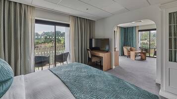 Habitación de hotel con cama, escritorio con televisor, balcón con sillas y vistas a zonas verdes.