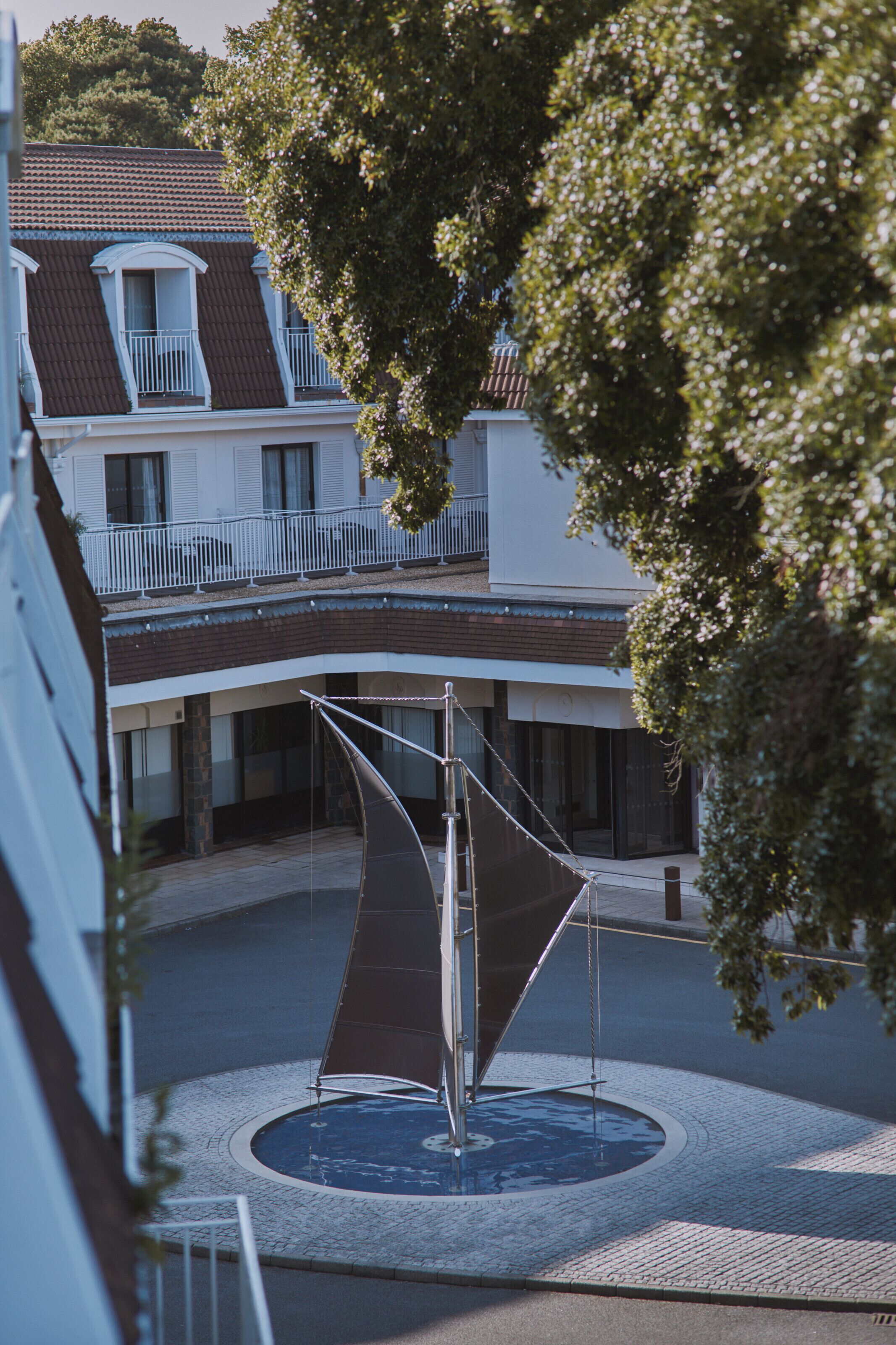 Une cour avec une structure ressemblant à une voile, un bâtiment doté d’un balcon et un arbre.
