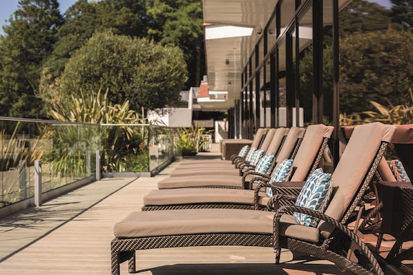 Una fila de sillones de mimbre con cojines, dispuestos en una terraza de madera con baranda de vidrio.