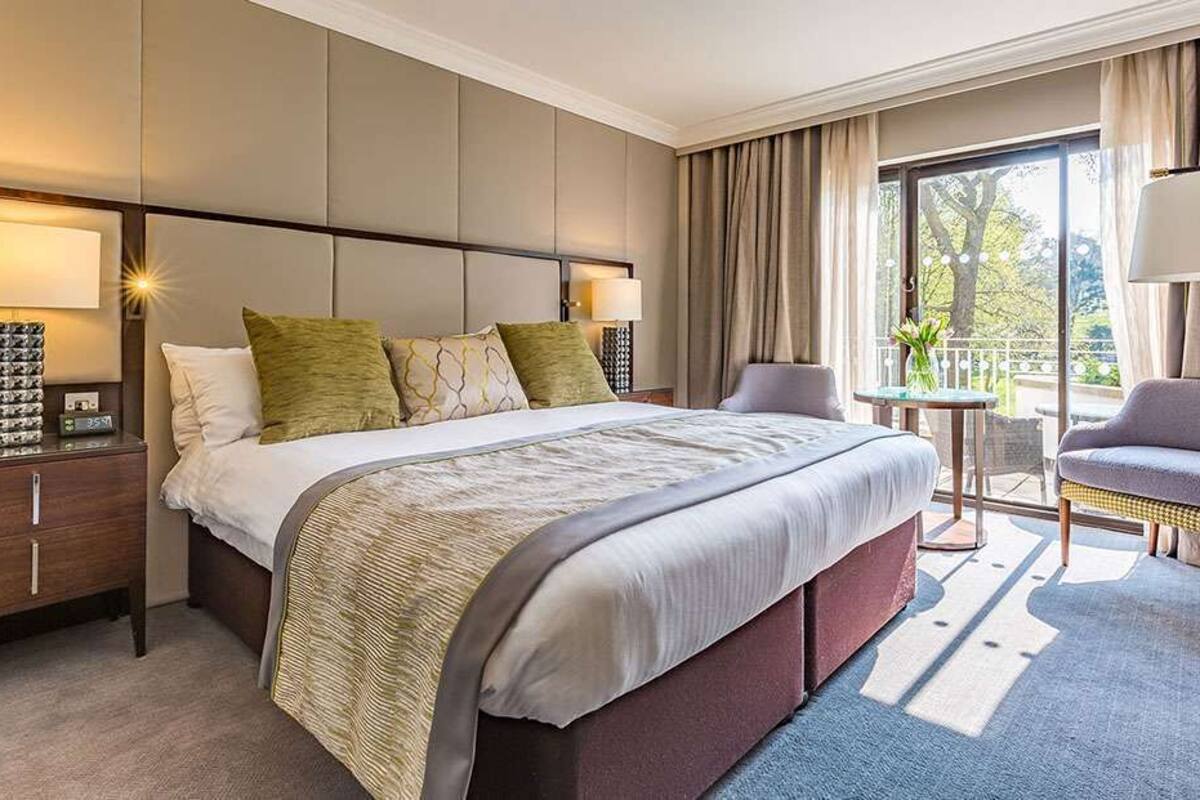 A hotel room with a large bed, a bedside table with a lamp, a chair, and a view of the outdoors.