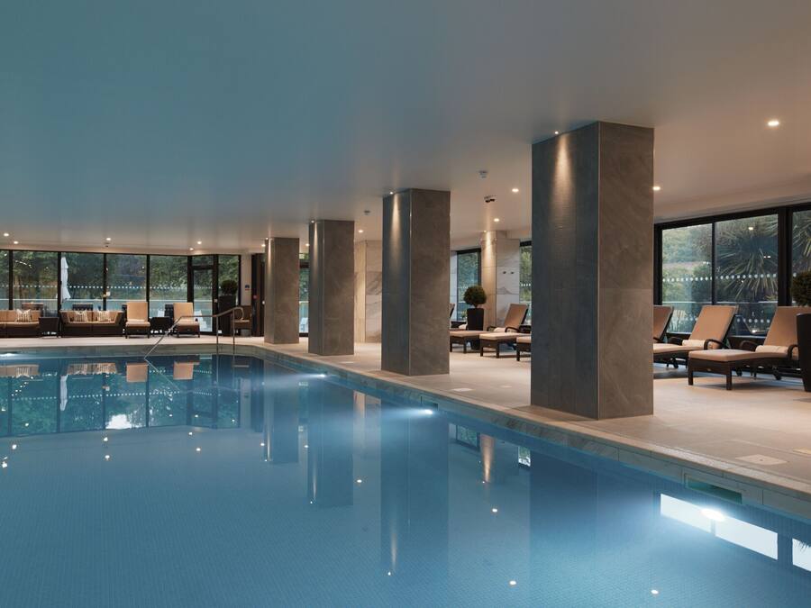 A modern indoor swimming pool with lounge chairs and a view of the outdoors.