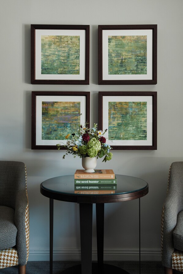 A room with four framed abstract paintings, a round table with a vase of flowers and books, and patterned chairs.