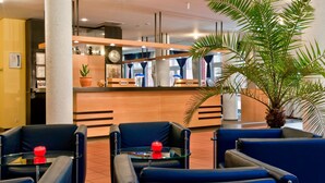 Un espace d’accueil d’hôtel comprenant un comptoir de réception, une horloge et des sièges, avec un objet décoratif rouge posé sur une table en verre.