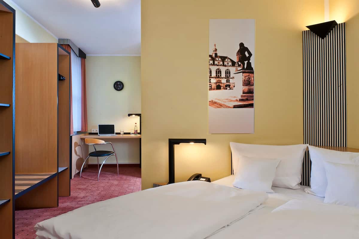 Een hotelkamer met een bed, bureau, stoel en een lampje aan de muur.