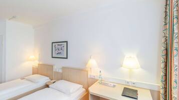 Una habitación de hotel con dos camas individuales, una mesita de noche con lámpara, un escritorio pequeño con lámpara y un cuadro enmarcado en la pared.