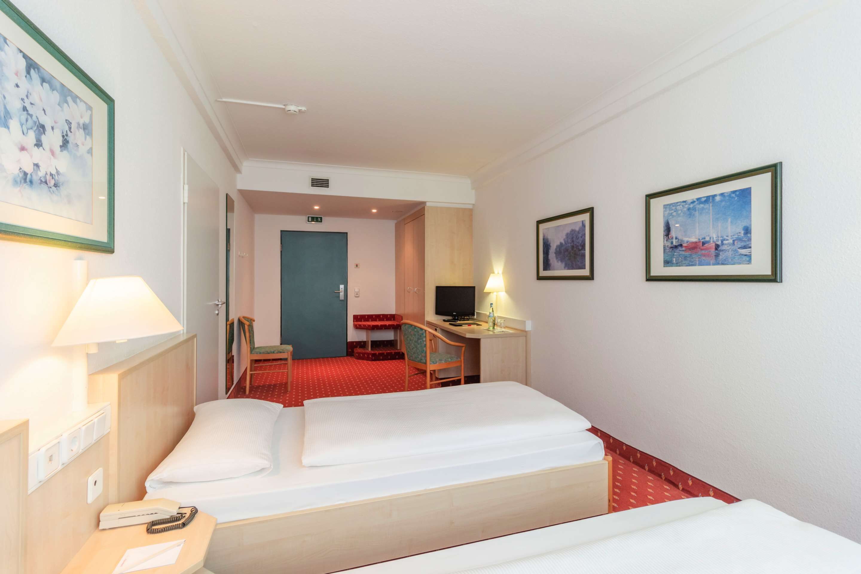 A hotel room with a bed, desk, chair, and two framed pictures on the wall.