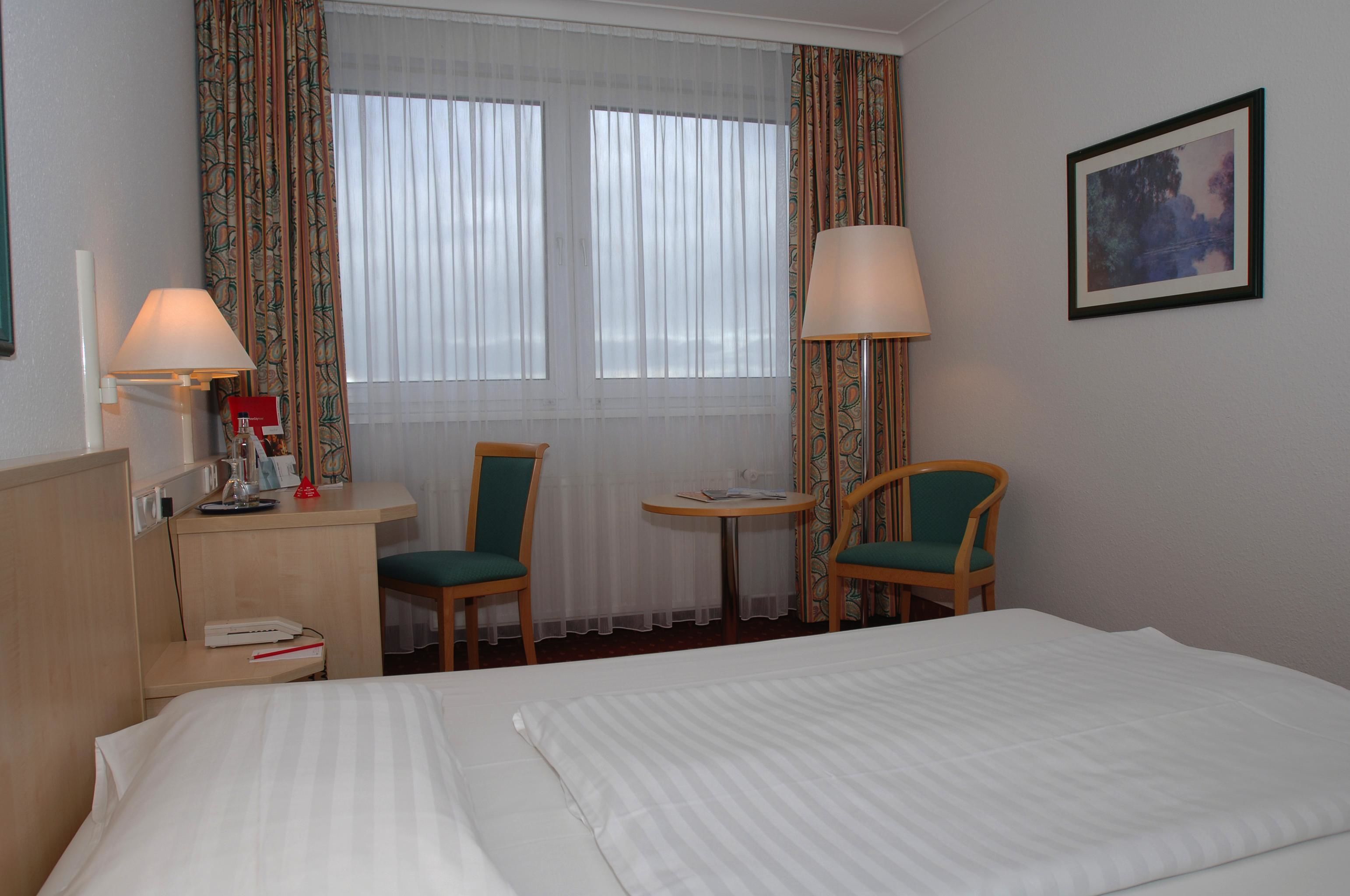 A hotel room with a bed, two chairs, a desk, a lamp, and a window with curtains.