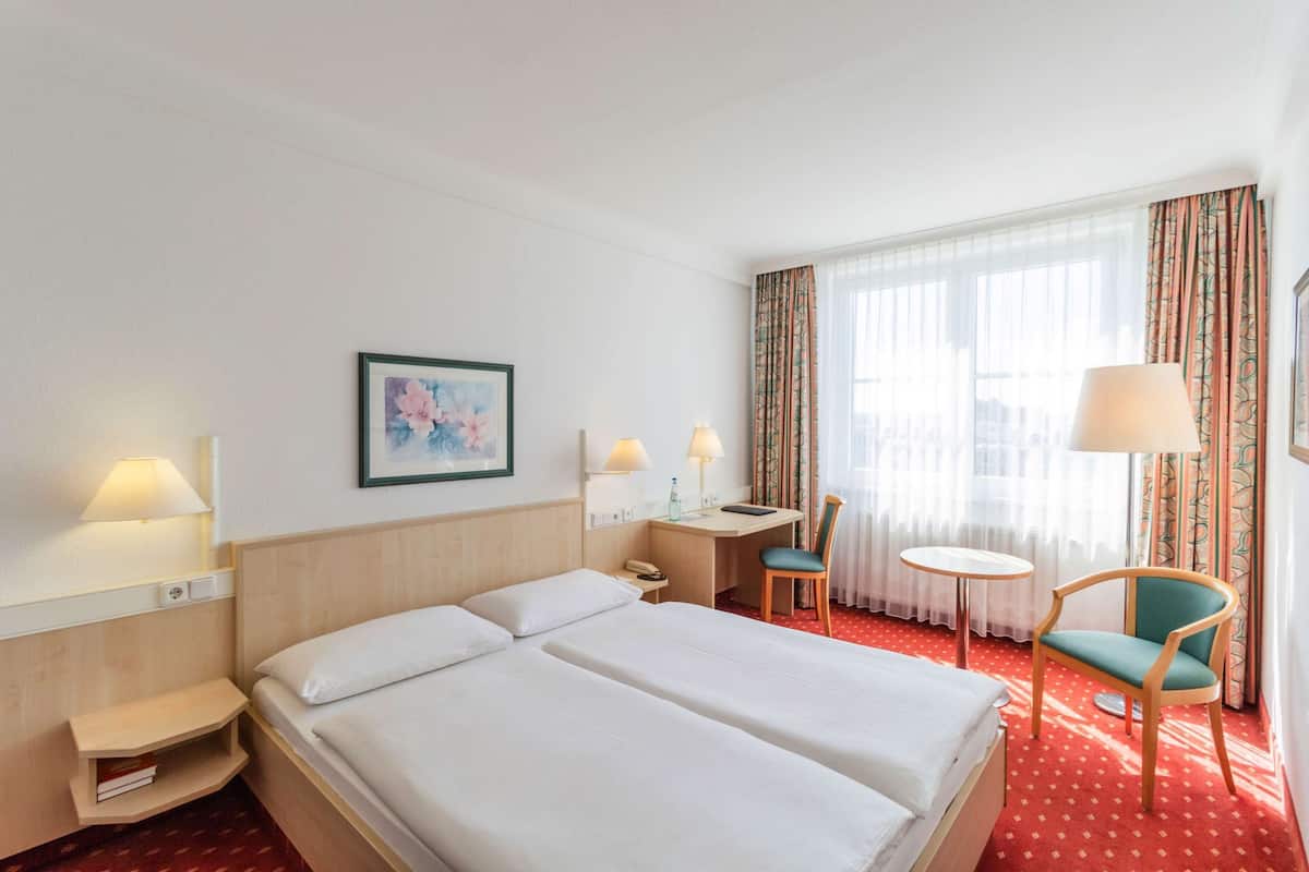 Une chambre d’hôtel avec un grand lit, un bureau, deux chaises et une fenêtre avec des rideaux.