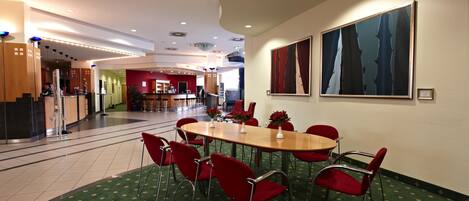 A hotel lobby with a reception desk, a dining area with a table and red chairs, and a large abstract painting on the wall.