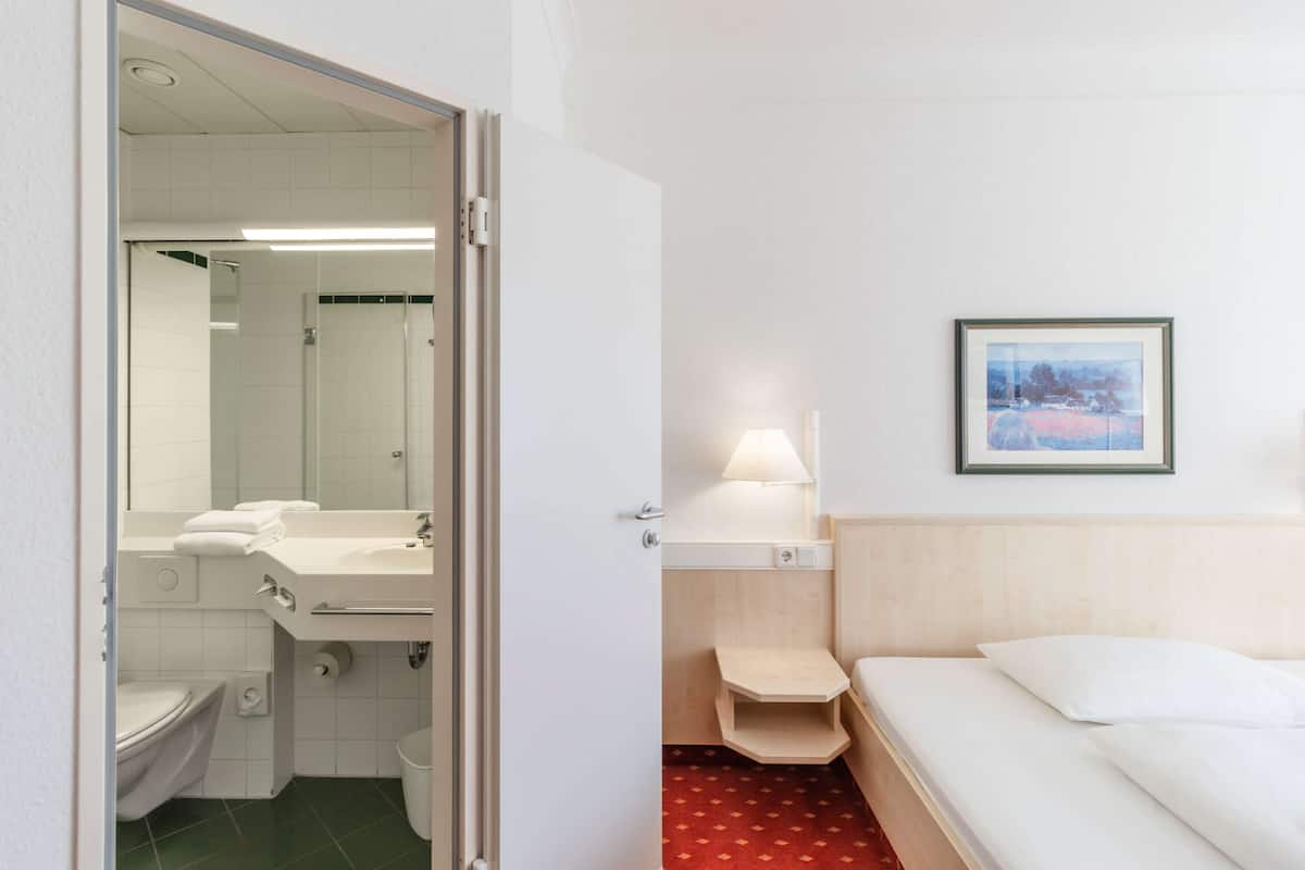 Une chambre d’hôtel avec un lit, une table de chevet avec une lampe, un tableau encadré au mur et une salle de bain visible à travers une porte ouverte.