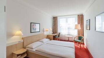 A hotel room with a large bed, bedside tables, a desk, two chairs, and a window with curtains.