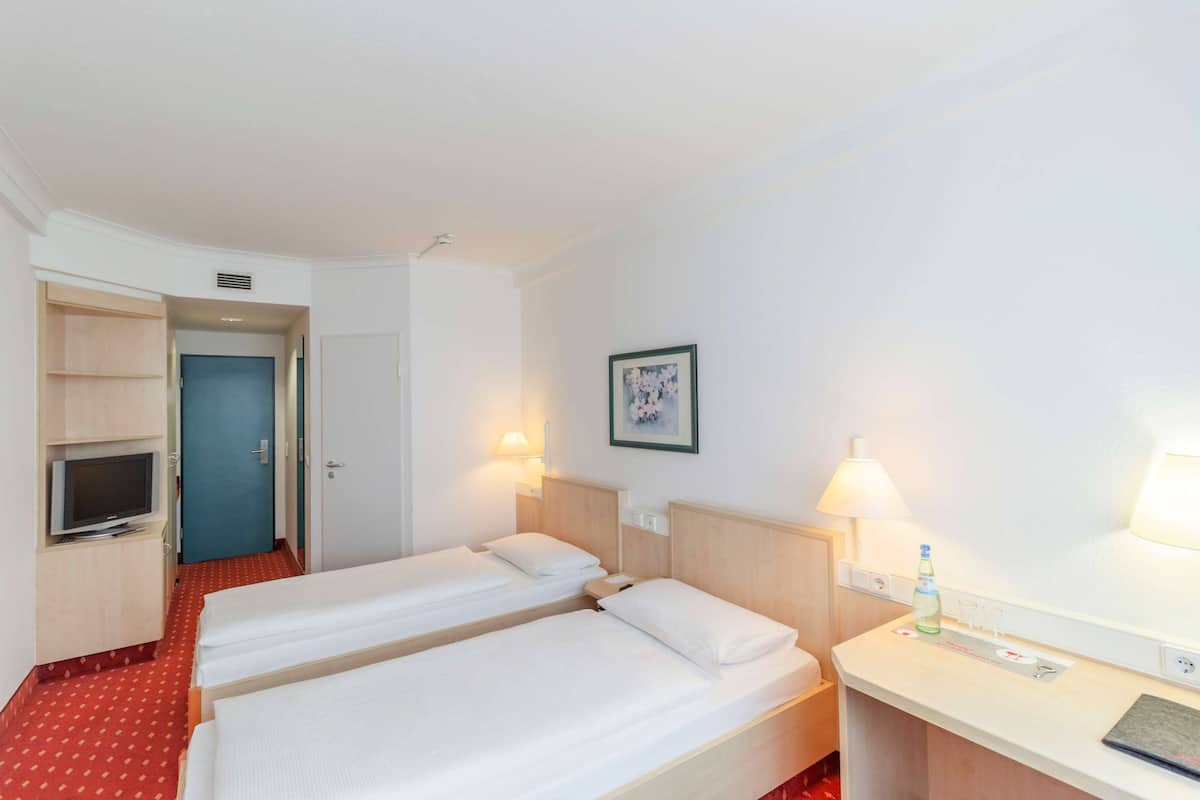 Une chambre d’hôtel avec deux lits simples, un bureau avec une lampe, une télévision et une porte donnant sur une salle de bain.