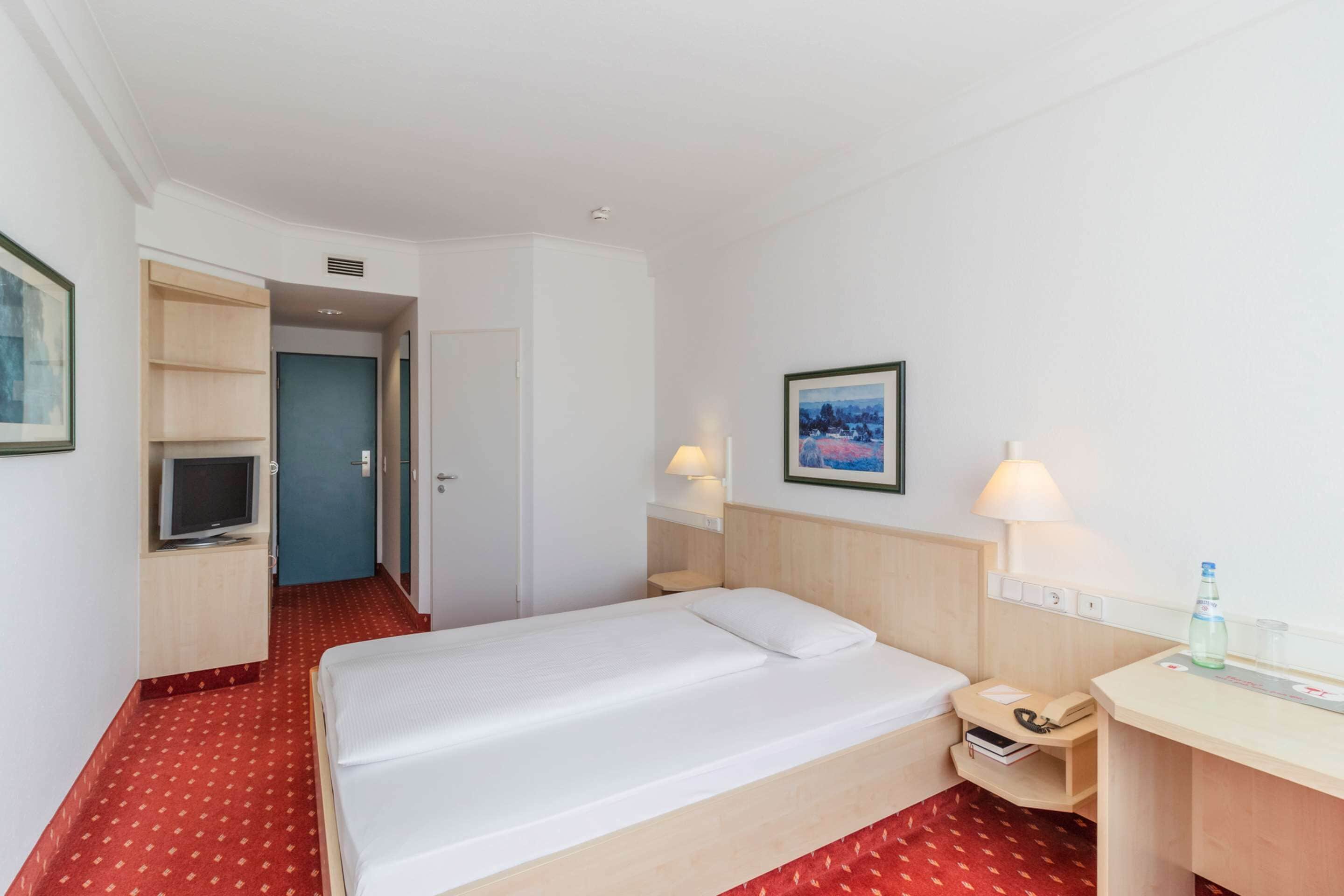 Una habitación de hotel con cama, mesita de noche, lámpara, televisor y escritorio con silla.