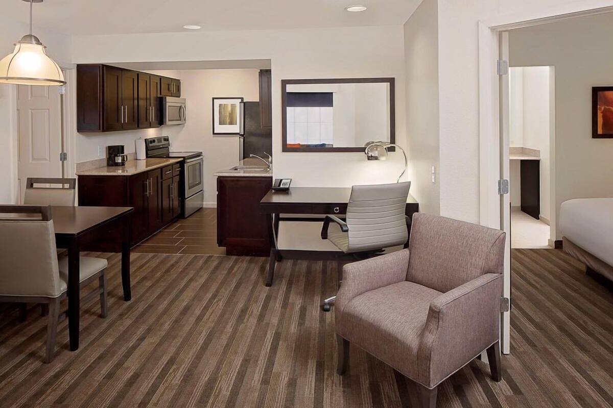 A compact hotel room with a kitchenette, a desk with a chair, a bed, and a large mirror.