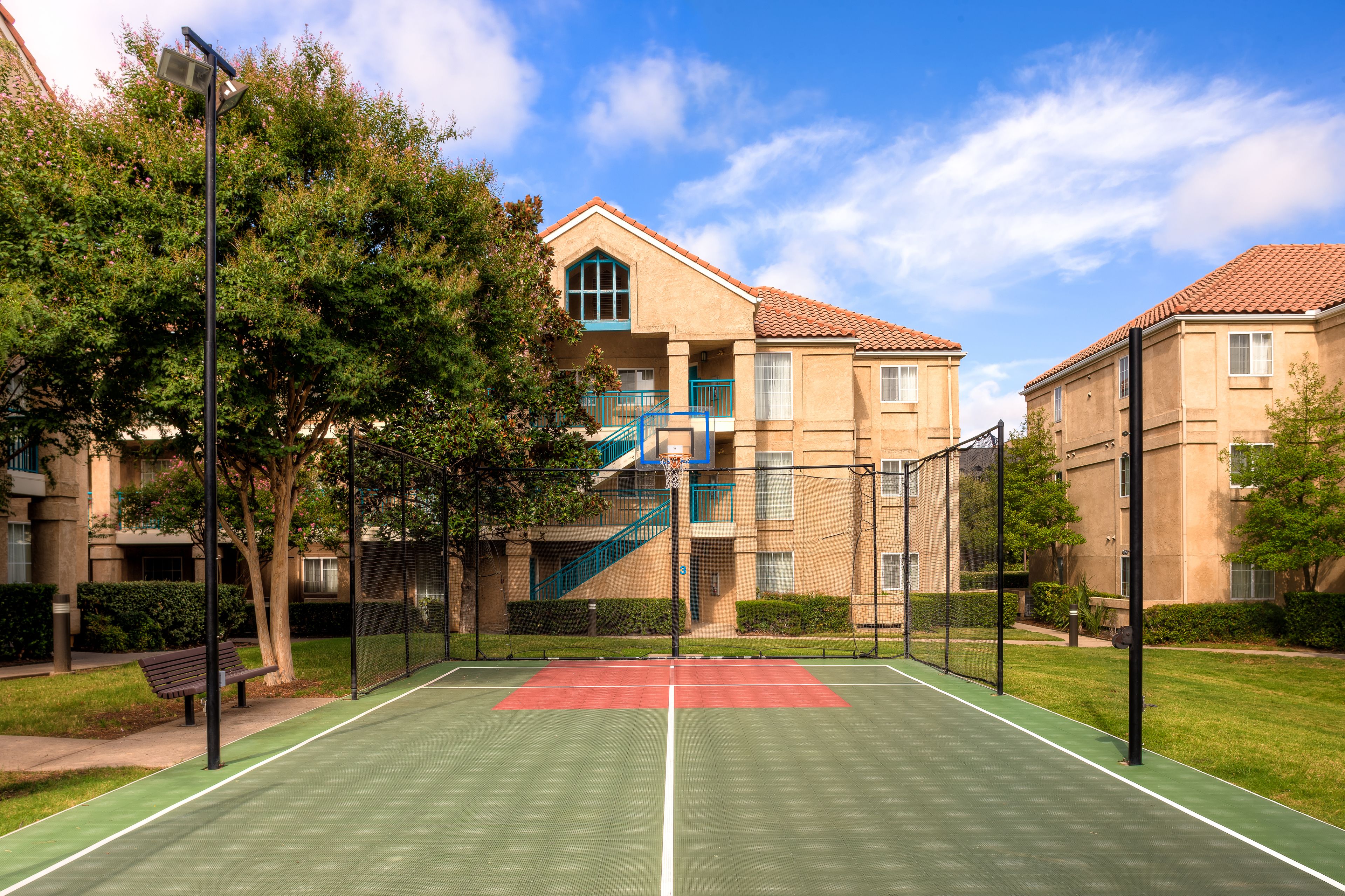 An outdoor tennis court with a net, surrounded by trees and a building with a red-tiled roof.