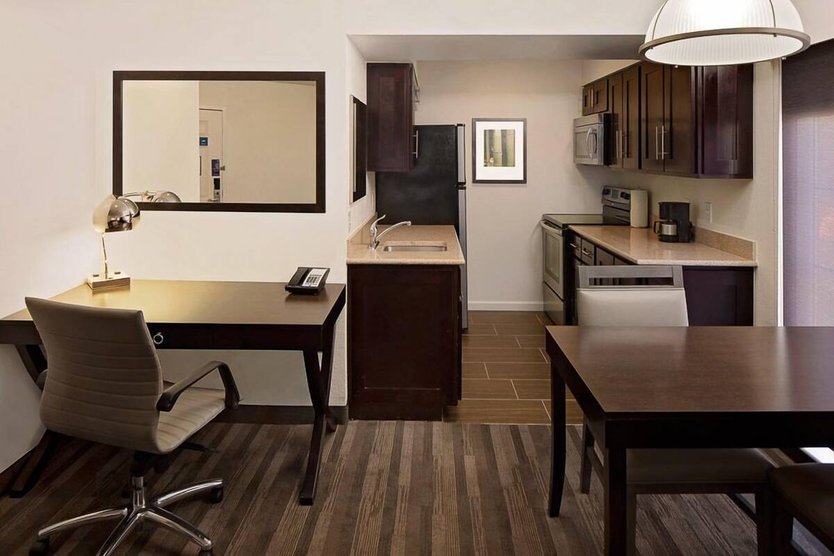 A compact hotel room with a kitchenette, a desk with a chair, a large mirror, and a dining table.
