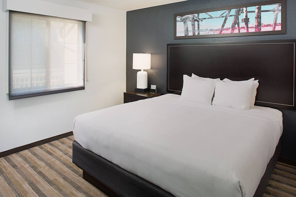 A hotel room with a large bed, a nightstand with a lamp, a window with blinds, and a framed picture on the wall.