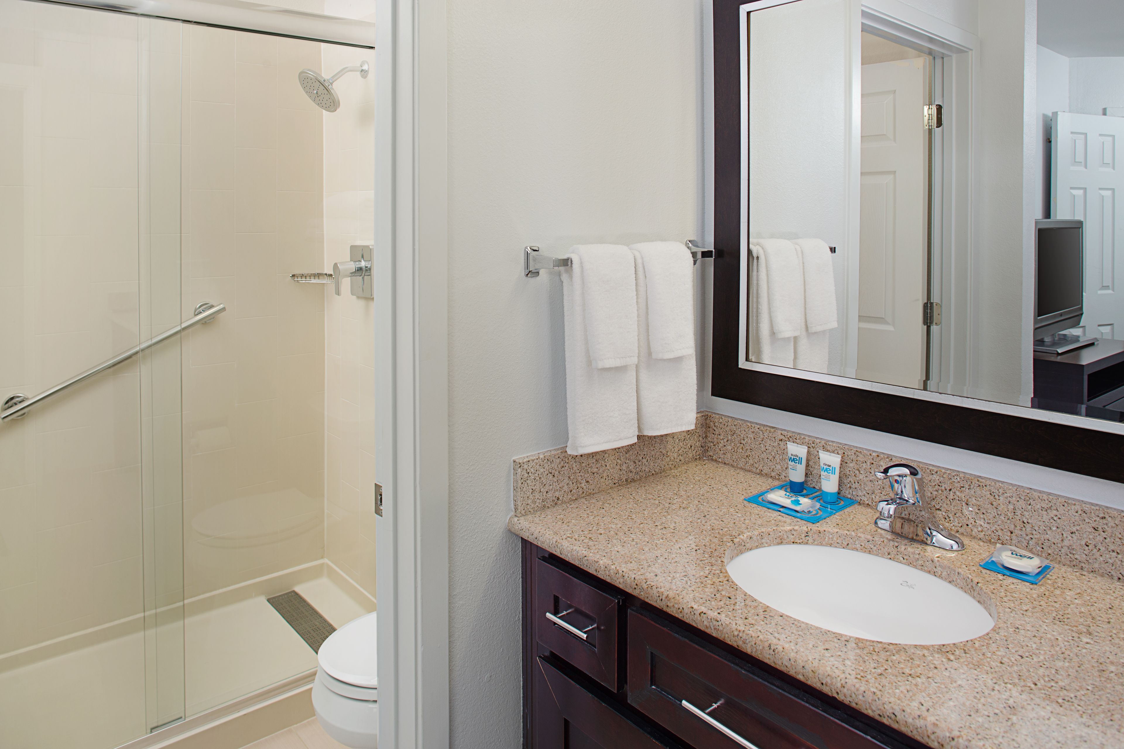 Un baño moderno con cabina de ducha de vidrio, un lavabo blanco con encimera de granito y un espejo grande sobre el lavabo.