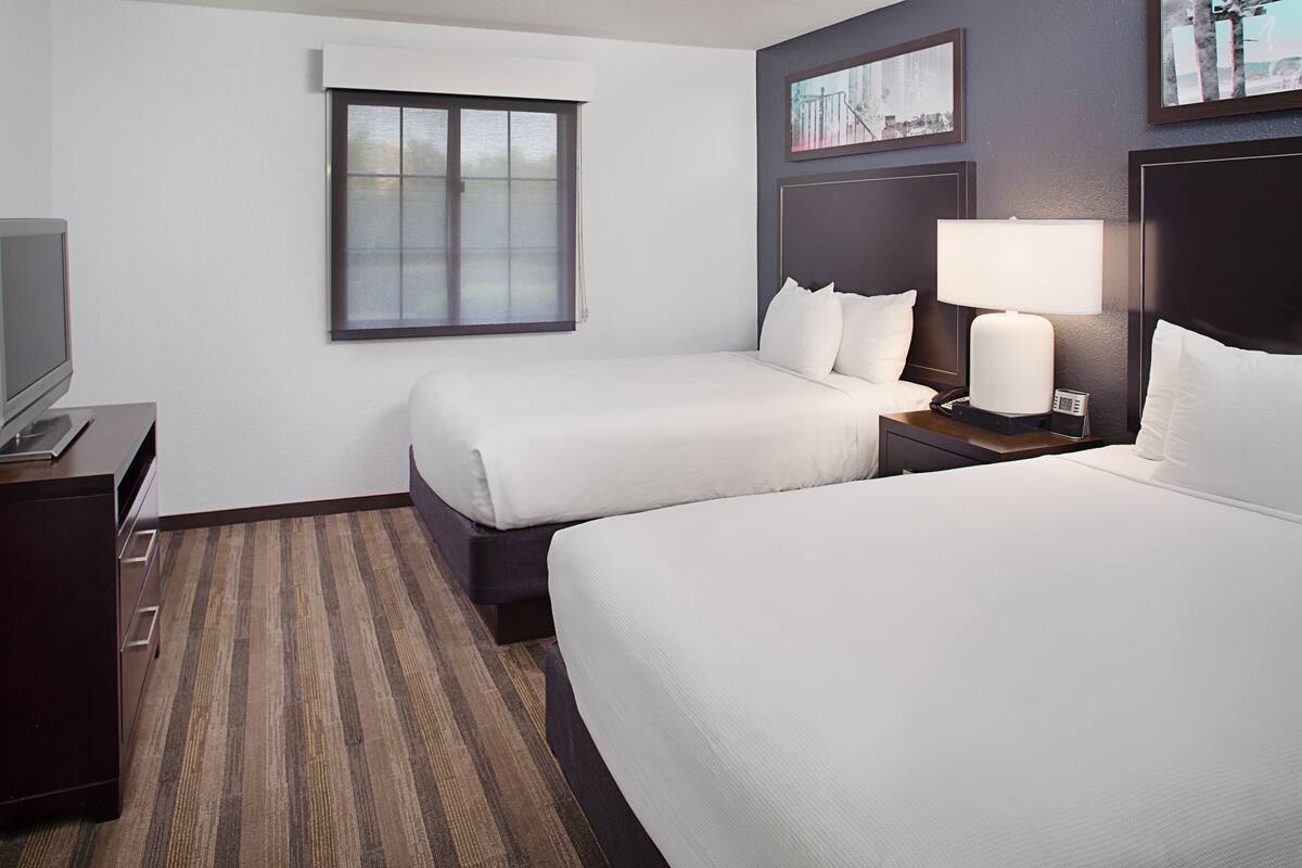 A hotel room with two beds, a desk with a television, a window, and a nightstand with a lamp.