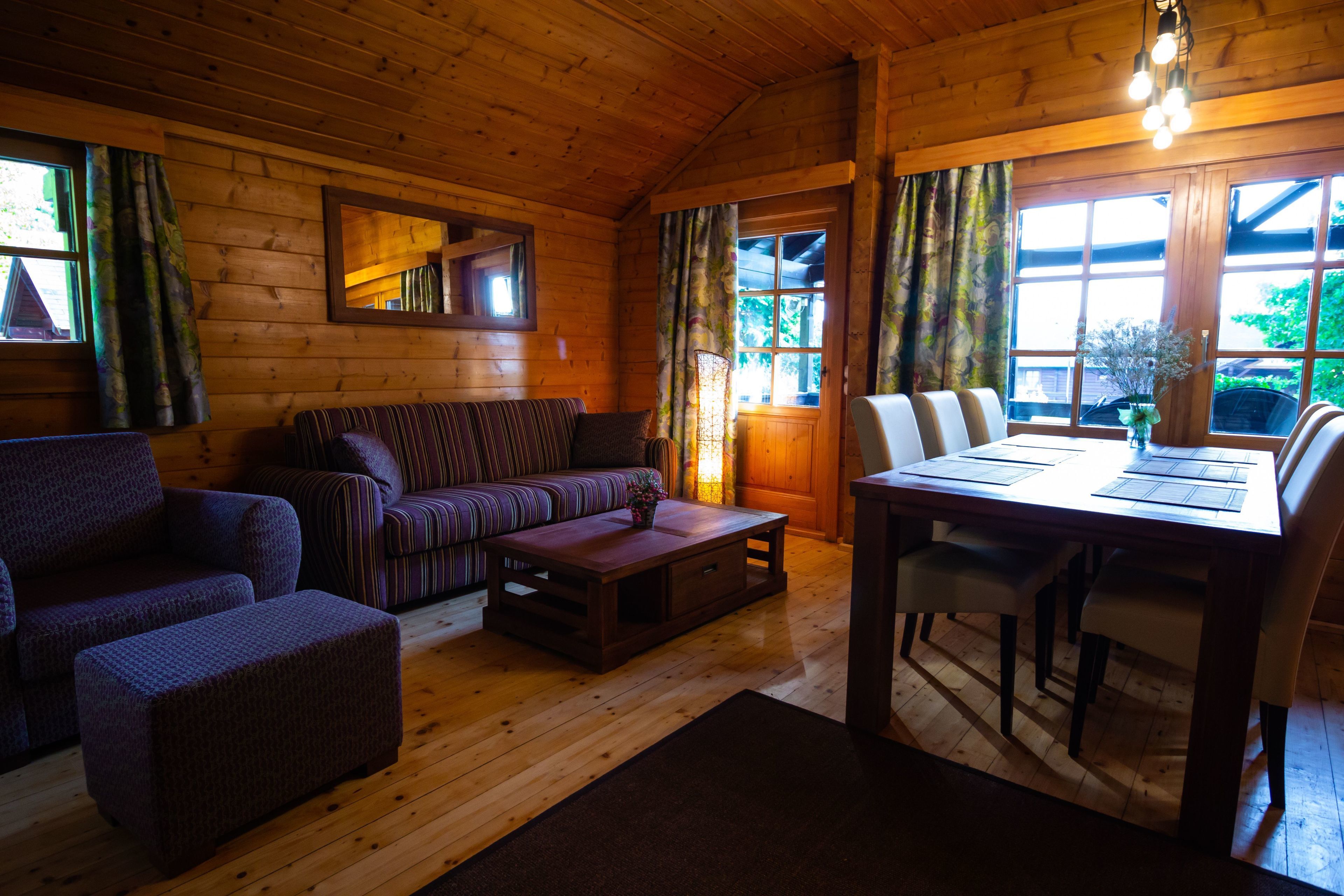 A cozy cabin interior with a wooden ceiling, a sofa, a dining table with chairs, and a window with curtains.