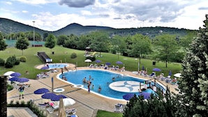 Piscina com várias pistas, rodeada por espreguiçadeiras e guarda-sóis, com colinas verdes e um céu nublado ao fundo.